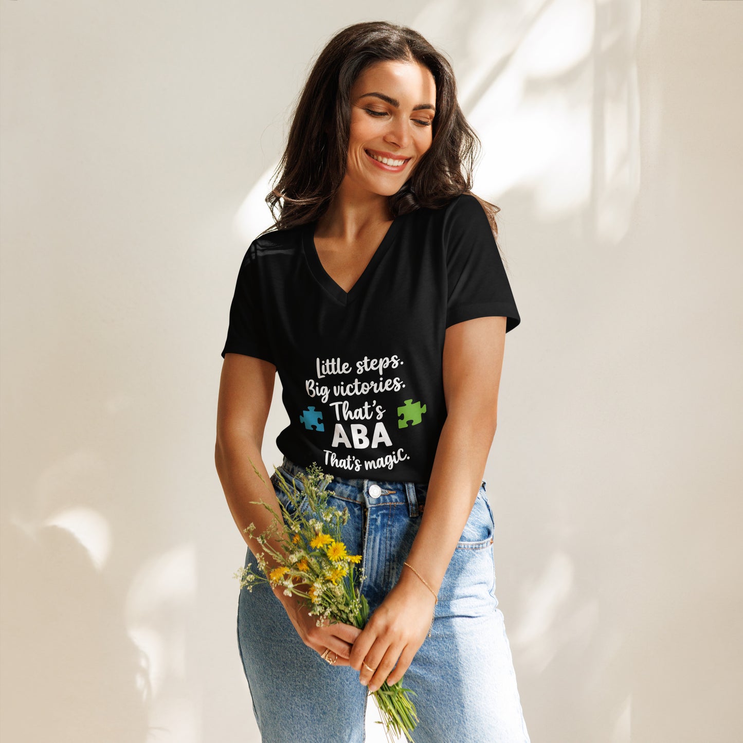 Woman wearing a black ABA therapy t-shirt that says ‘Little Steps, Big Victories – That’s ABA Magic,’ featuring colorful puzzle pieces – relaxed v-neck shirt for RBTs and ABA professionals.